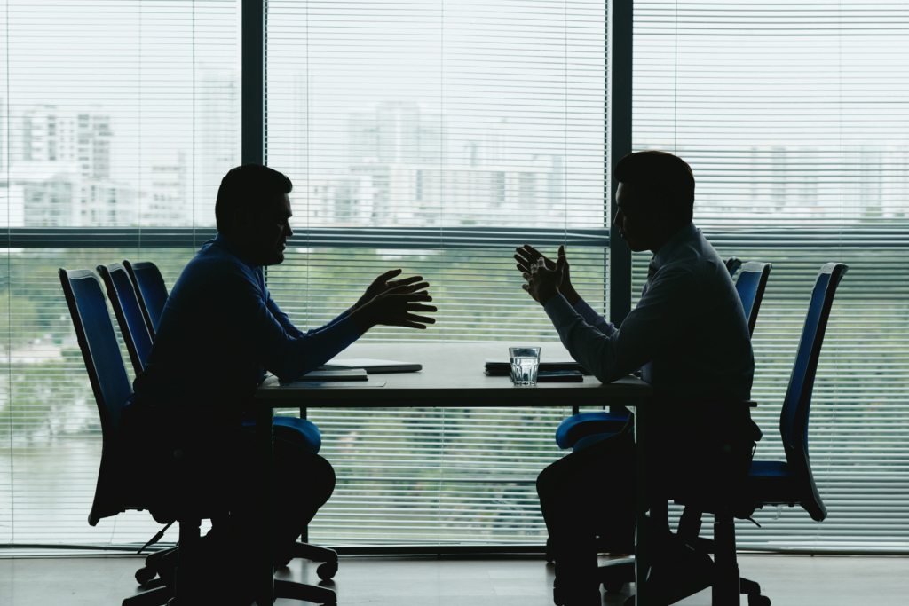 two-human-outlines-against-shuttered-office-window-sitting-opposite-each-other-negotiating