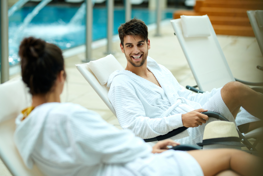 young-couple-bathrobes-talking-while-sitting-by-pool-spending-weekend-spa-focus-is-happy-man